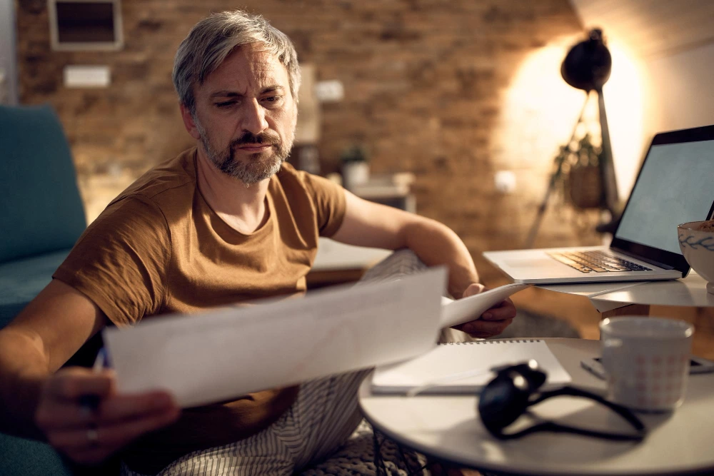 A middle-aged man in casual clothes reviews documents at a home desk