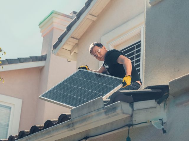 A male technician wearing safety gear installing a solar panel on a roof.